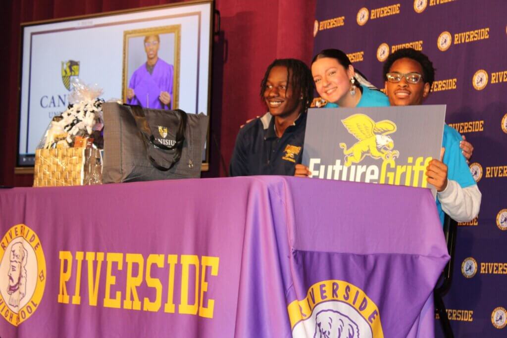 Three students pose behind a Riverside table with a Future Griff sign; a display screen and gift basket are visible in the background.