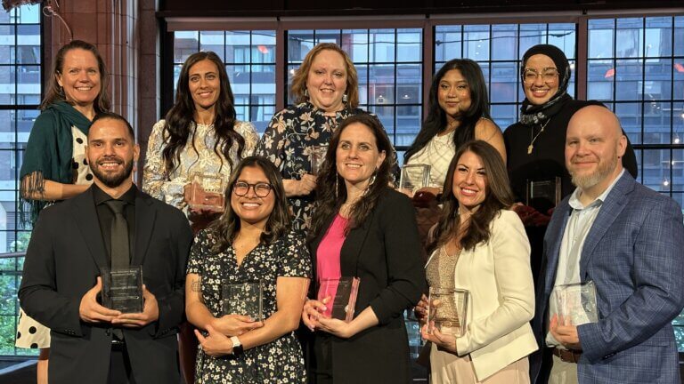 Eleven people posing indoors, each holding a glass award and smiling at the camera, with large windows and a cityscape visible in the background.