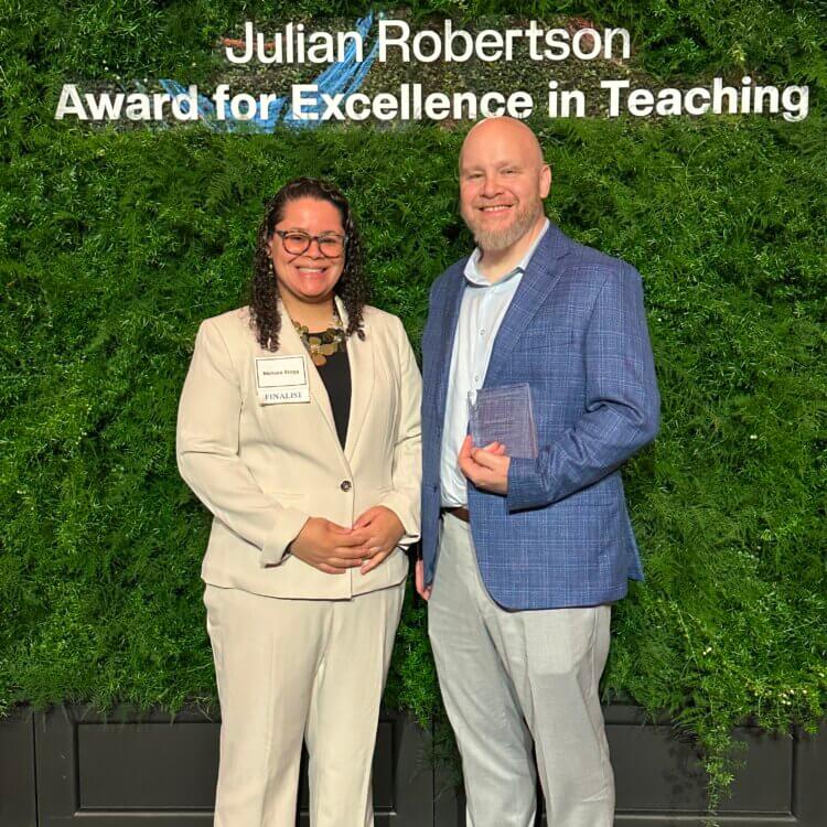 Two people in business attire stand in front of a green backdrop with the sign Julian Robertson Award for Excellence in Teaching. One holds a glass award.