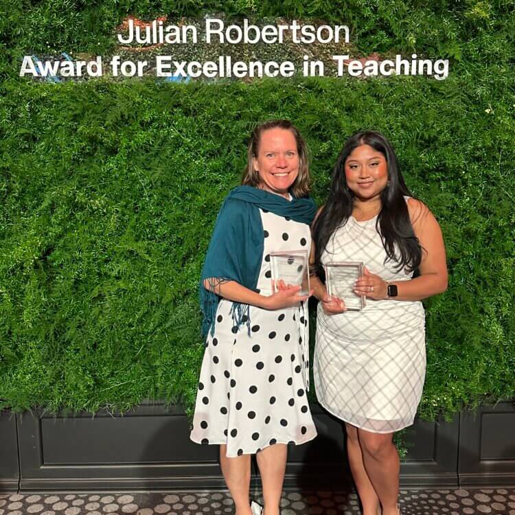 Two women stand in front of a green backdrop holding awards beneath a sign that reads Julian Robertson Award for Excellence in Teaching.