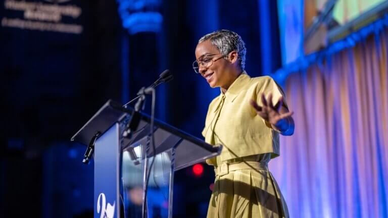 Tony Award winner and alumna Kara Young, dressed in a yellow outfit, speaks at a podium with a microphone in a brightly lit event space.