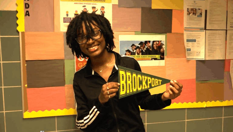 A student wearing glasses and a dark jacket, holds a SUNY Brockport pennant and smiles