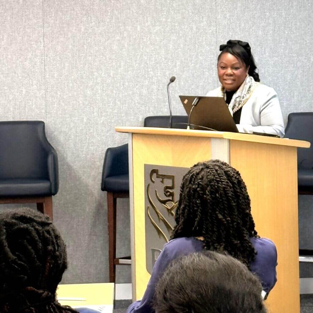 A woman stands at a podium with a laptop, speaking to an audience in a conference room