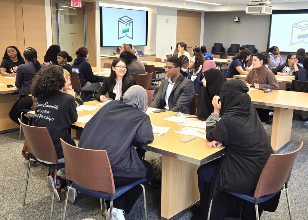 Students and professionals sit in groups at tables in a modern classroom, engaged in discussion.