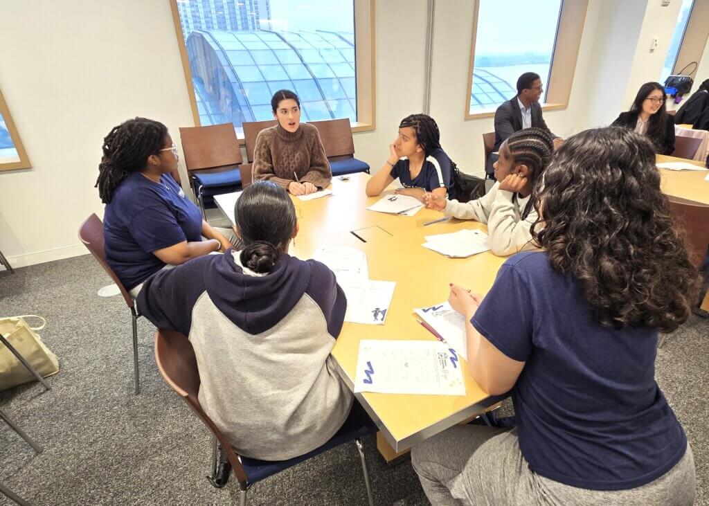 A group of students sit around a table with papers, talking and listening during a discussion in a meeting room.