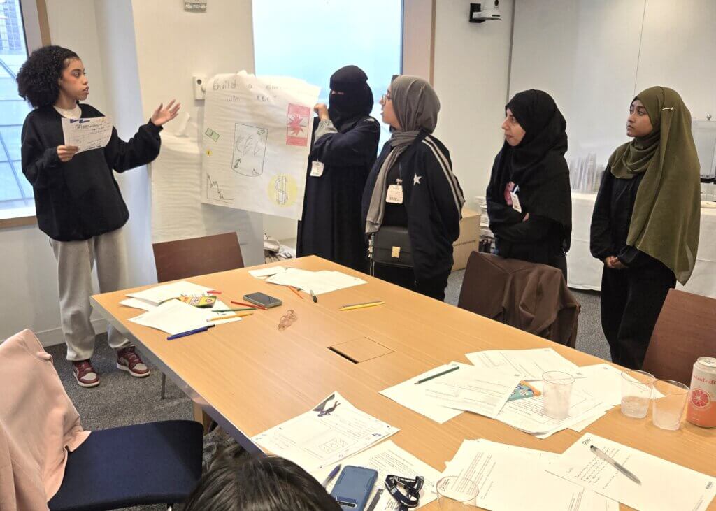 Five young women stand at the front of a room; one presents while others hold a poster.