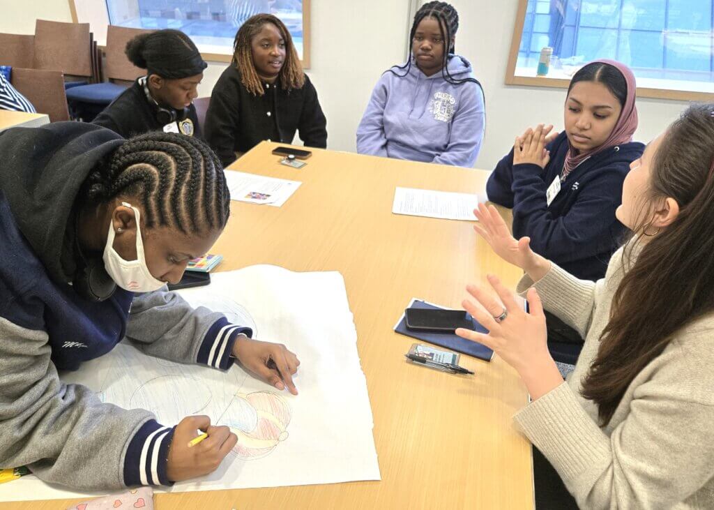 Six young women sit around a table; one draws on a large sheet of paper while the others talk and listen.