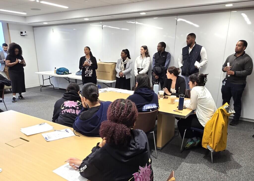 A group of people stands at the front of a conference room, speaking to attendees seated at tables