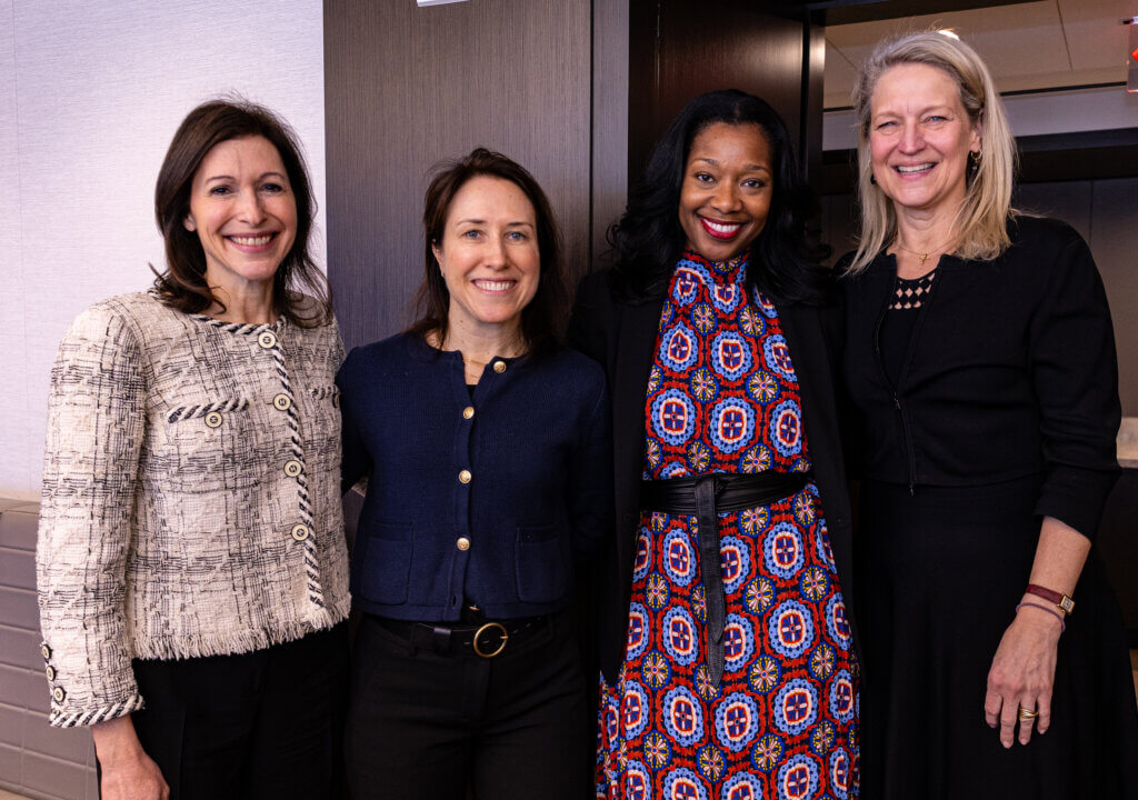 Four women standing side by side indoors, smiling at the camera; they are dressed in business or professional attire.
