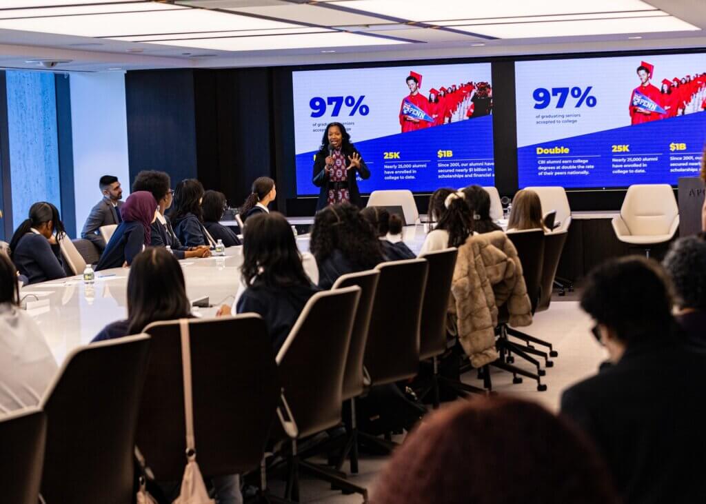 A woman speaks to an audience in a conference room with two screens behind her displaying a statistic: 97% of graduating seniors.