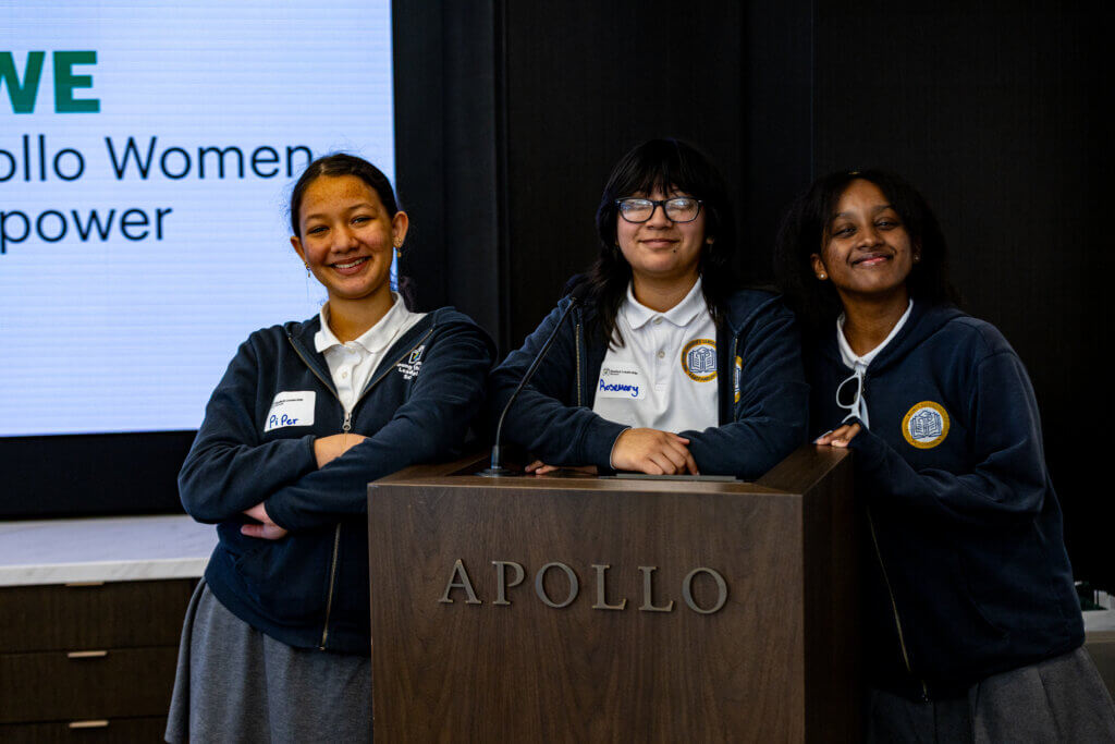 Three students in school uniforms stand and smile at a podium labeled APOLLO with a screen in the background displaying partial text.
