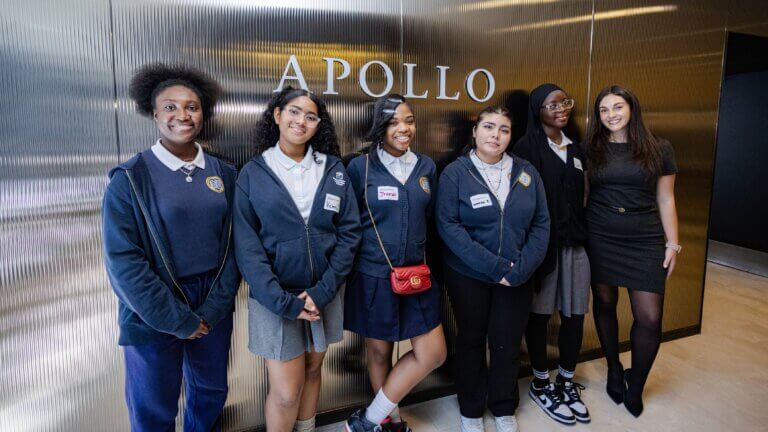 Six young women, five in school uniforms and one in business attire, stand and smile in front of a wall with the word APOLLO displayed.
