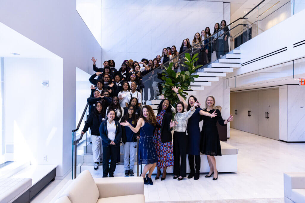 A large group of people, mostly women, pose and smile on and around a staircase in a modern, well-lit building.