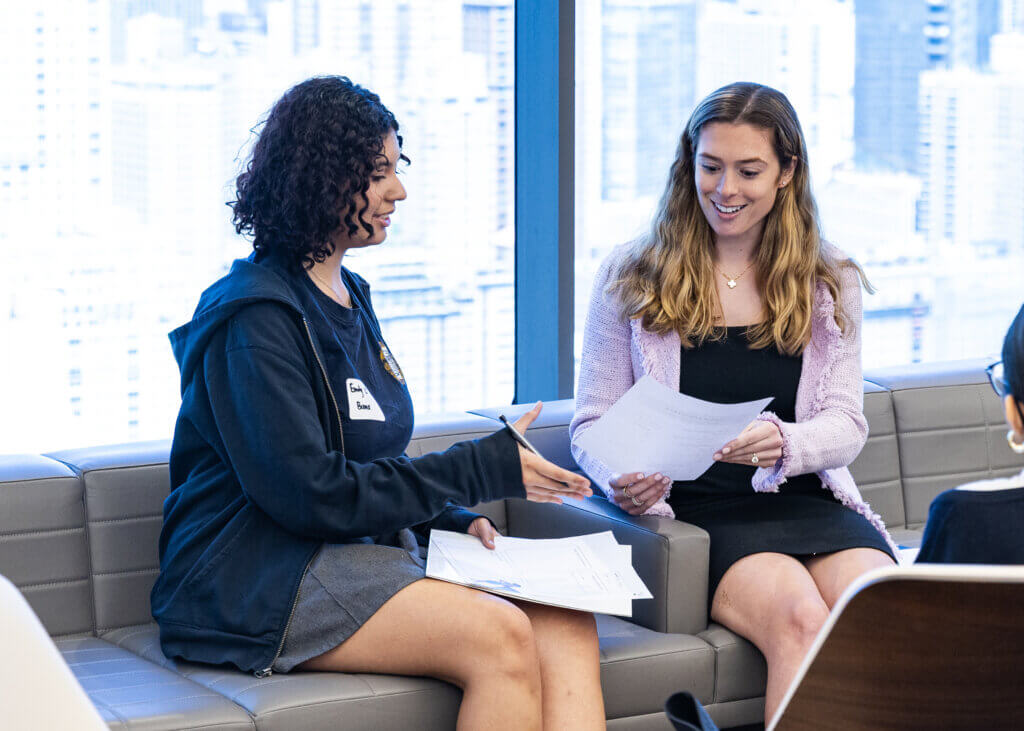Two women sit on a couch by a window, holding papers and having a discussion in an office setting with a cityscape in the background.
