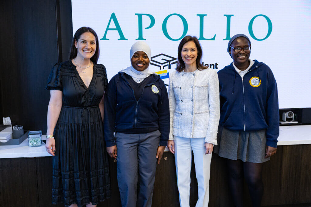 Four women stand side by side, smiling, in front of a screen displaying the word APOLLO. Two are in school uniforms, and two are in professional attire.