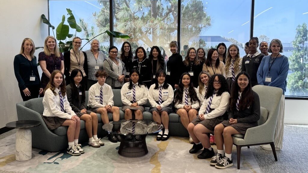 A group of students in uniforms and adults pose together in an office lounge area with large windows, plants, and modern furniture.