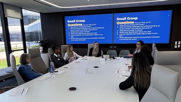 Six people sit around a conference table in a modern meeting room, with discussion questions displayed on two large screens at the front.