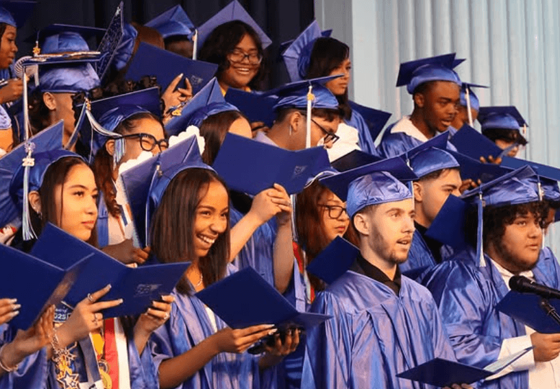 A group of students in blue caps and gowns hold diplomas and smile during a graduation ceremony.