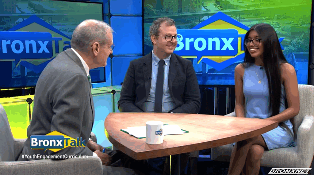 Three people sit at a round table having a discussion on the set of BronxTalk, with the show’s logo visible on monitors in the background.