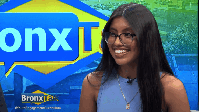 A woman with long dark hair and glasses smiles during a BronxTalk TV interview, with the show’s logo visible in the background.