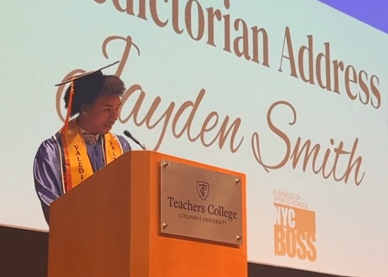 A student in a cap and gown speaks at a podium marked Teachers College Columbia University during a valedictorian address on college decision day, with Jayden Smith displayed on the screen behind.
