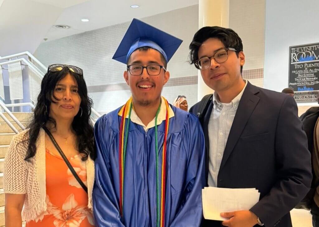 Three people pose indoors; the center person wears a blue graduation cap and gown with multicolored cords, flanked by a woman in a floral dress and a man in a suit holding papers.