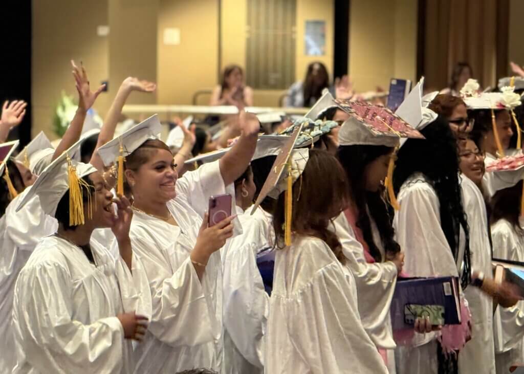 A group of graduates in white caps and gowns stand together, some smiling, waving, and holding phones during a graduation ceremony.