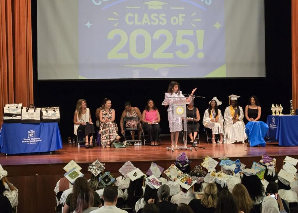 A woman speaks at a podium on stage during a graduation ceremony, with graduates in caps and gowns seated behind her and a Class of 2025! sign displayed above.