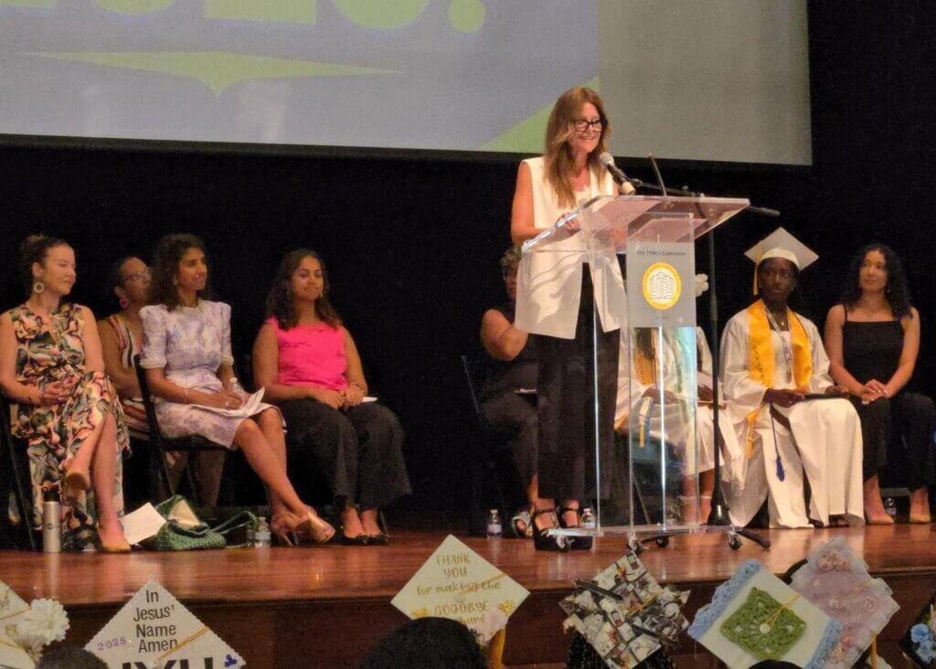 A woman speaks at a podium on a stage during a graduation ceremony, while graduates and attendees sit behind her.