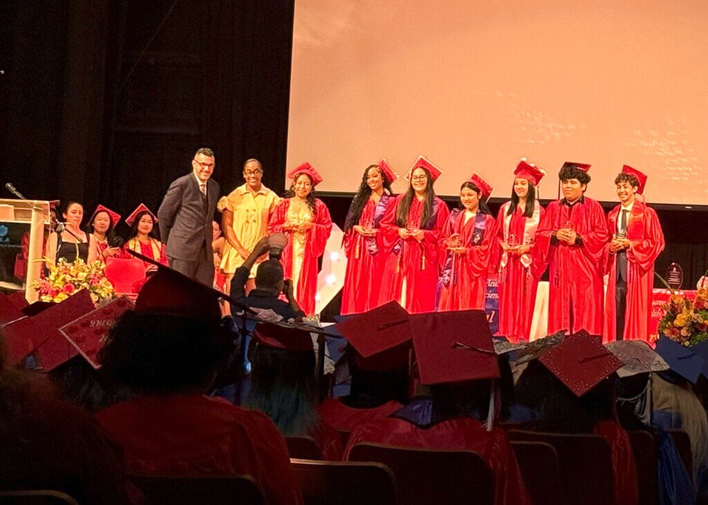 A group of students in red caps and gowns stand on stage during a graduation ceremony, with an adult and an audience visible.