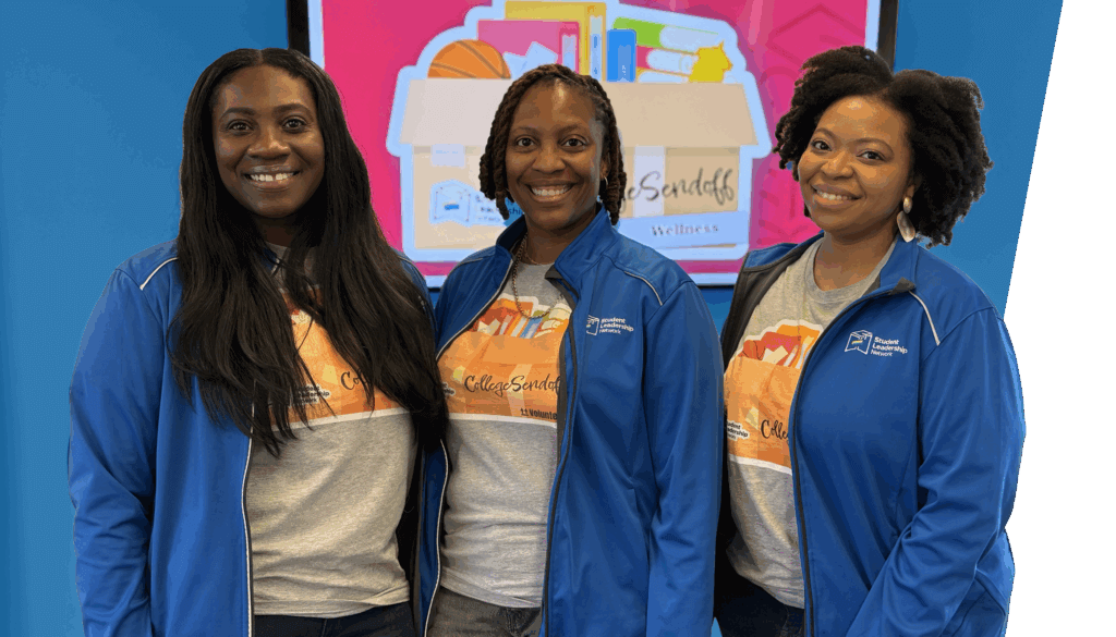 Three women wearing matching blue jackets and themed T-shirts stand smiling in front of a screen displaying a colorful wellness box graphic.