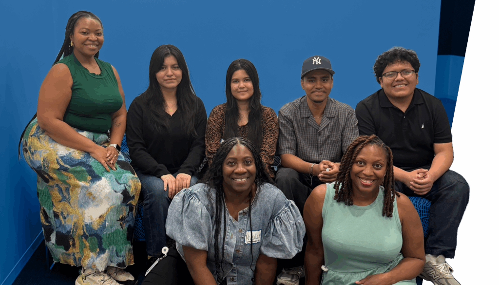 Seven people pose together indoors, with four sitting on a couch and three seated on the floor in front. They are casually dressed and smiling at the camera.