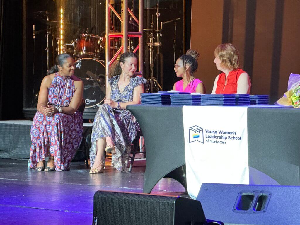 Four women sit on stage near a table with a Young Womens Leadership School of Manhattan banner, with stacked folders and a bouquet of flowers on the table.
