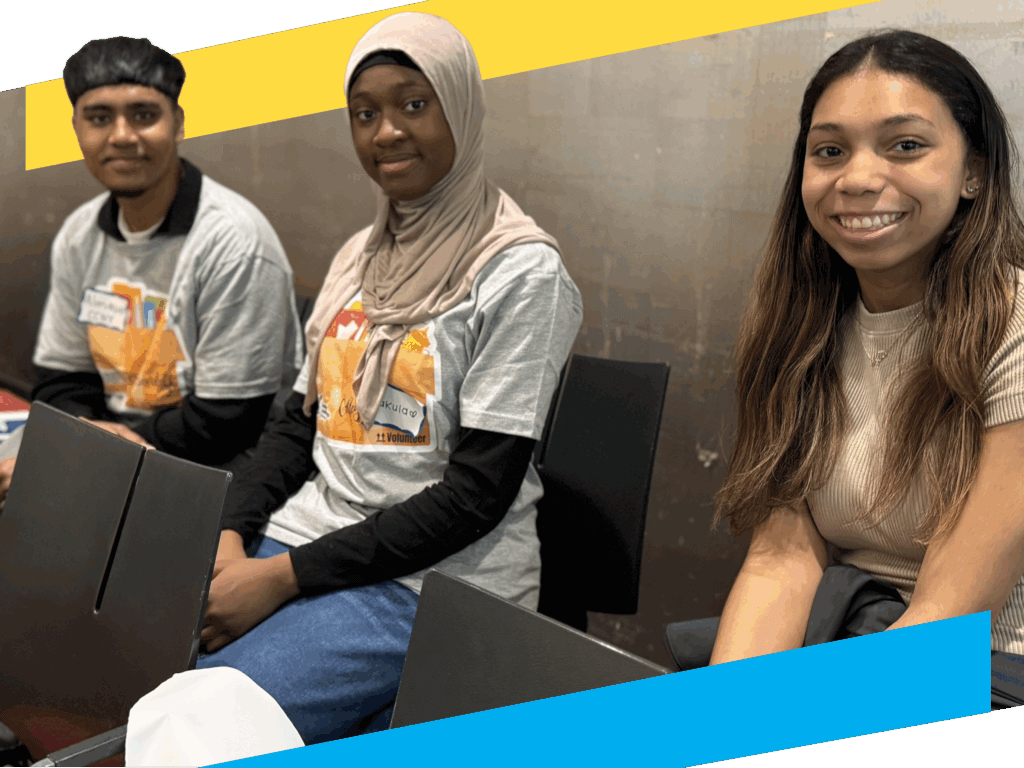 Three young people sitting indoors, two wearing gray event t-shirts and one in a beige top, all facing the camera and smiling.