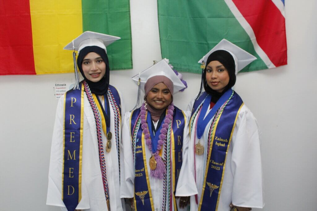 Three graduates in white gowns and hijabs celebrate college decision day, standing before colorful flags with PREMED stoles, medals, and cords, smiling proudly at the camera.