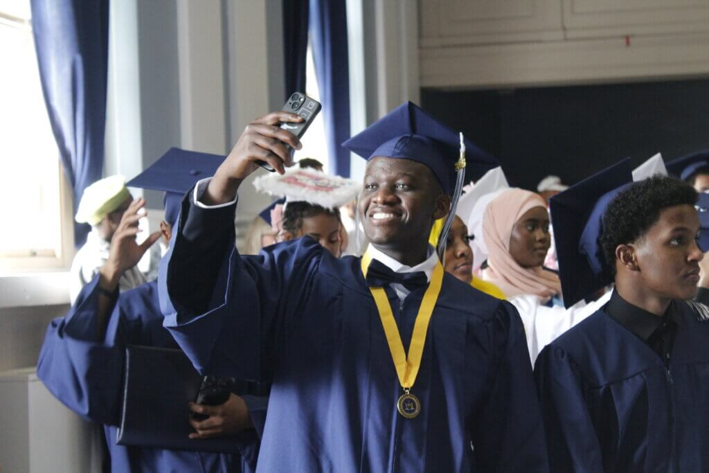 A graduate in a navy cap and gown smiles while taking a selfie, surrounded by other graduates in similar attire at a commencement ceremony.