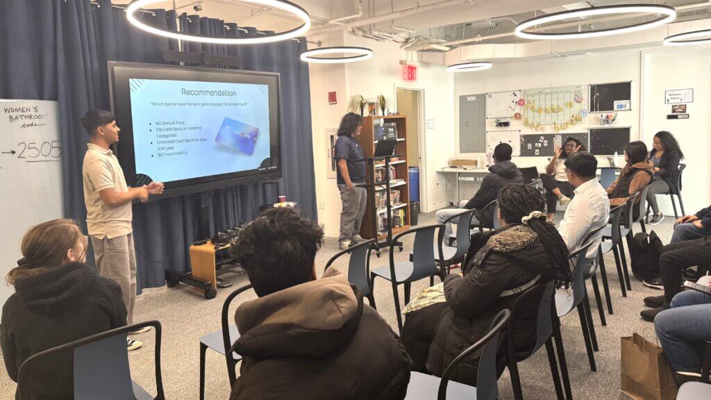A group of students sits in a classroom, listening to two presenters standing by a screen displaying a slide titled Recommendation with personal finance tips.