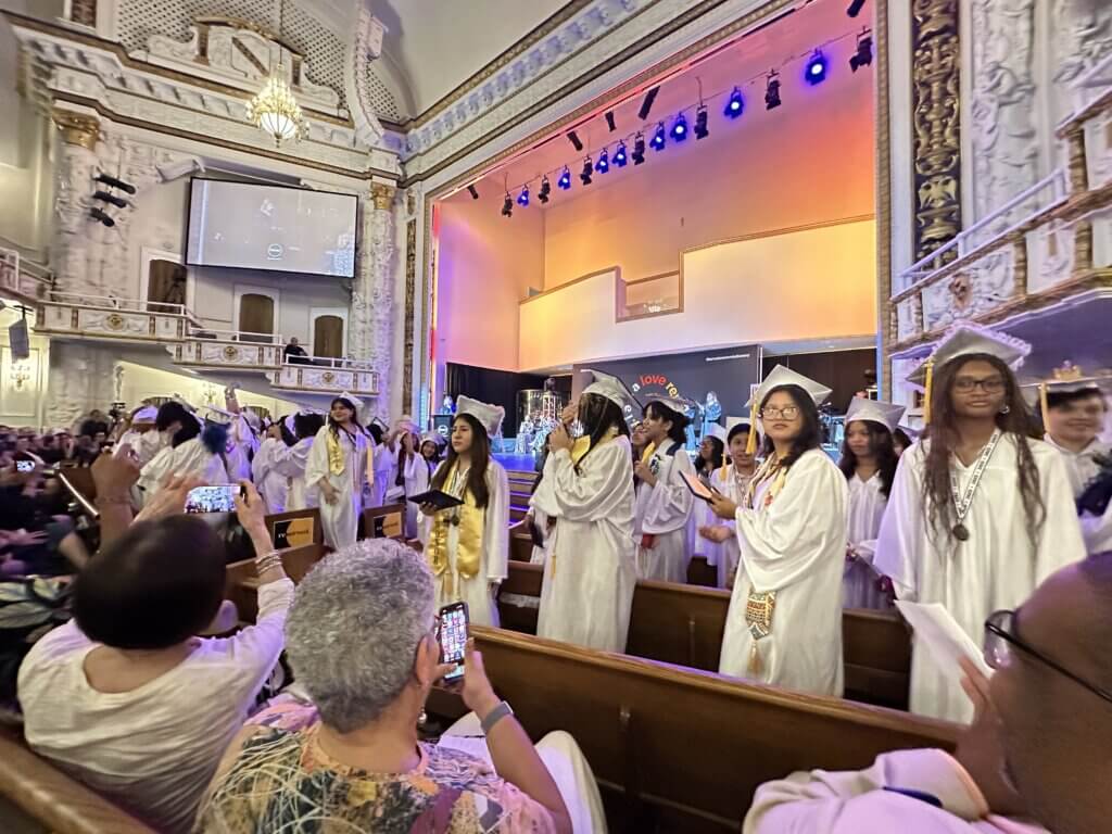 Graduates in white caps and gowns walk down the aisle of an ornate theater while audience members take photos, capturing a memorable college decision day during the graduation ceremony.