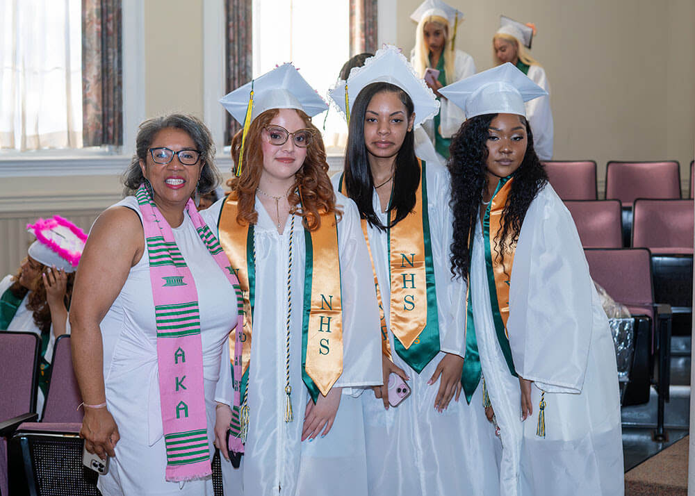 A woman in academic regalia stands at a podium while two graduates in caps and gowns stand beside her, smiling, during a college decision day commencement ceremony.