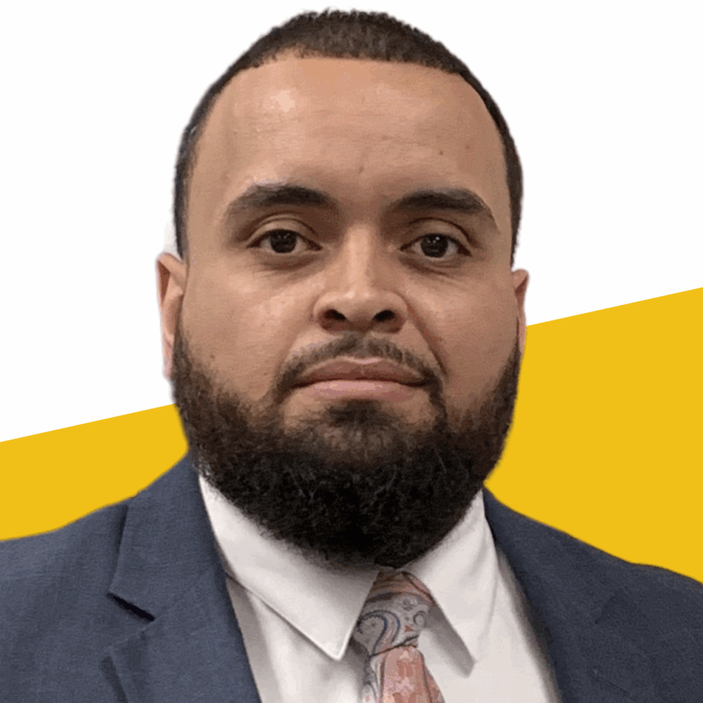 A man with a trimmed beard in a blue suit, white shirt, and patterned tie, poses confidently in front of a white and yellow background.