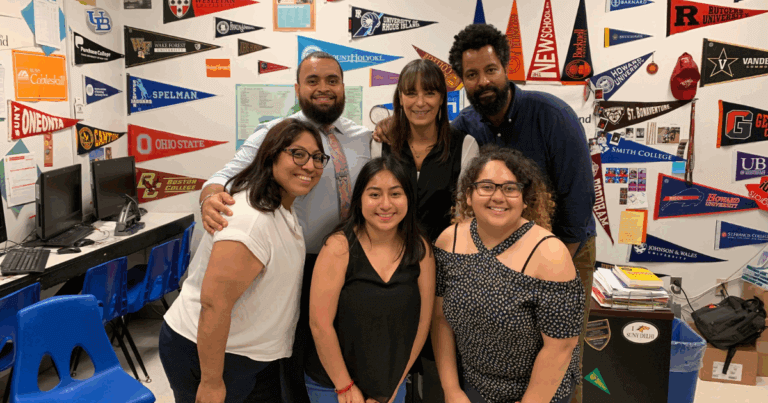 Six adults, including Benjamin Ramos, smiling and posing together in a classroom with college pennants covering the wall behind them.