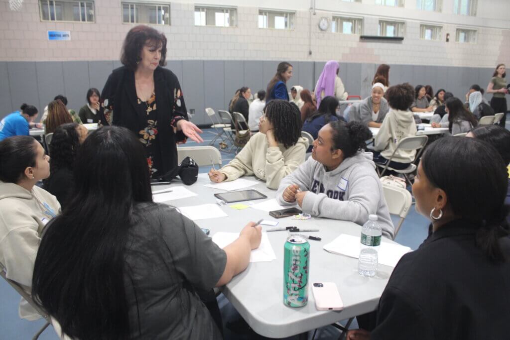 A woman stands and speaks to a group of seated people at a table in a large room, with papers, drinks, and writing materials on the table.