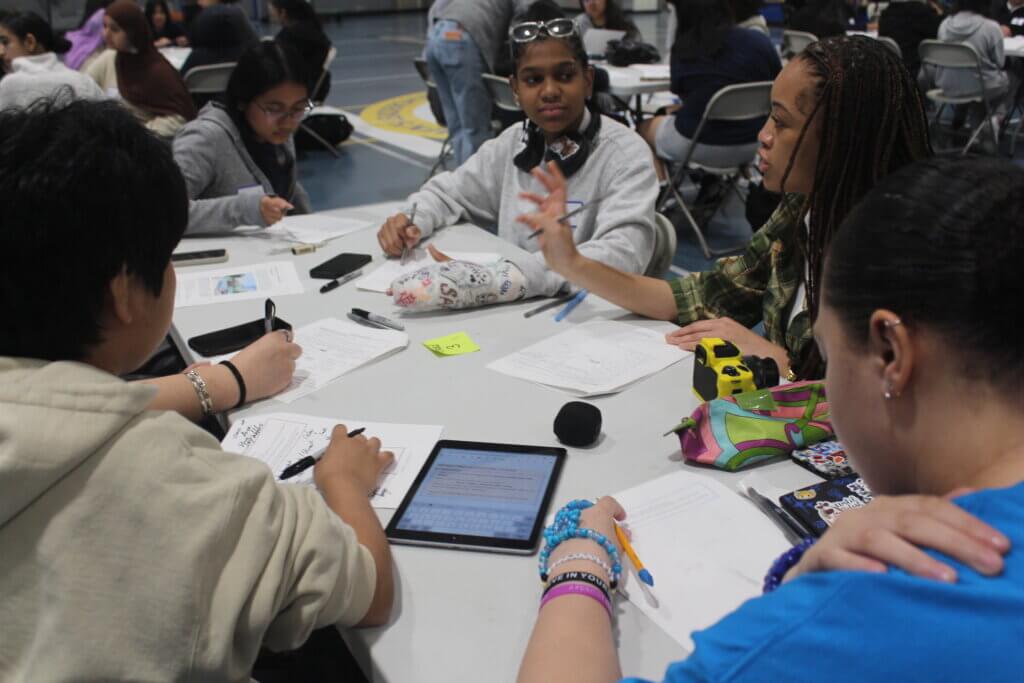 A group of students sit around a table working on papers and tablets, discussing and writing in a gymnasium setting.
