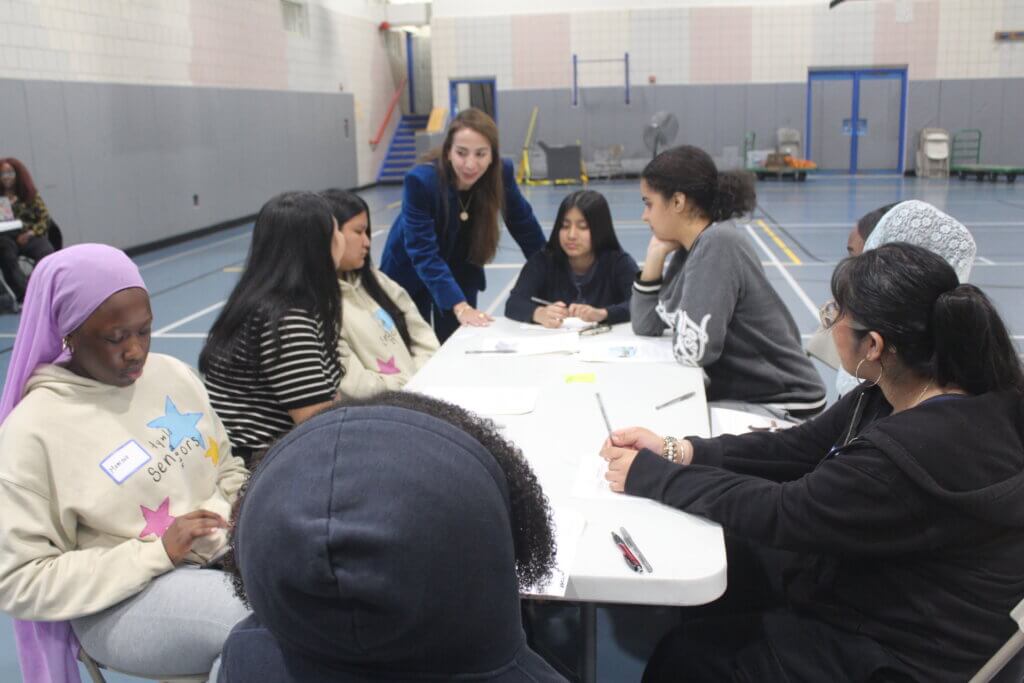 A group of people sit around tables in a gymnasium, engaged in discussion. A woman stands, addressing the group, while others listen and write notes.