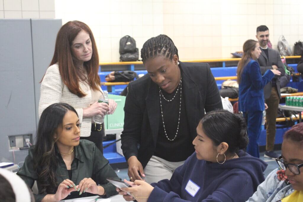 Four women are gathered around a table in conversation, with one woman standing and pointing at a phone.