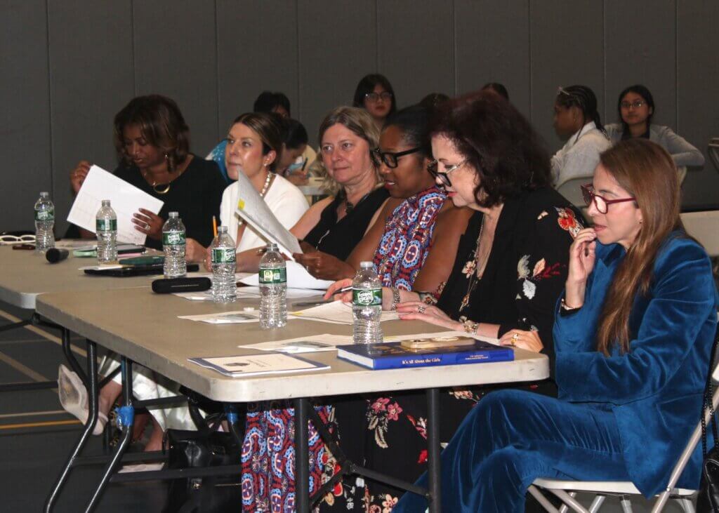 Six women sit at a table with papers, judging an event in a gymnasium with people seated in the background.