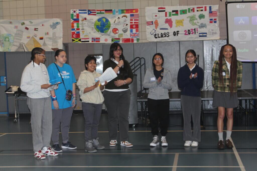 Seven students stand in a gym, some holding papers and microphones