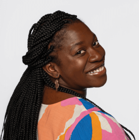A woman with long braided hair smiles over her shoulder, wearing a colorful patterned top and gold earrings against a plain white background.