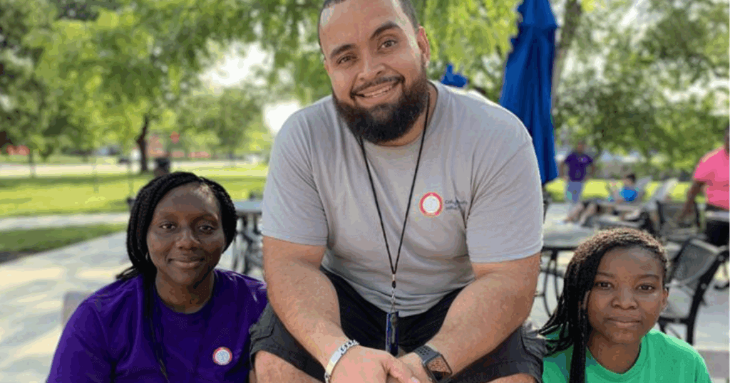 Benjamin crouches between two students as they all smile at the camera on a sunny day.