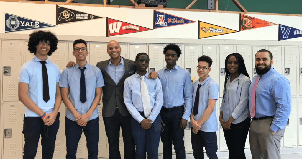 Eight people stand in front of lockers, dressed in business casual attire, with college pennants displayed on the wall behind them.